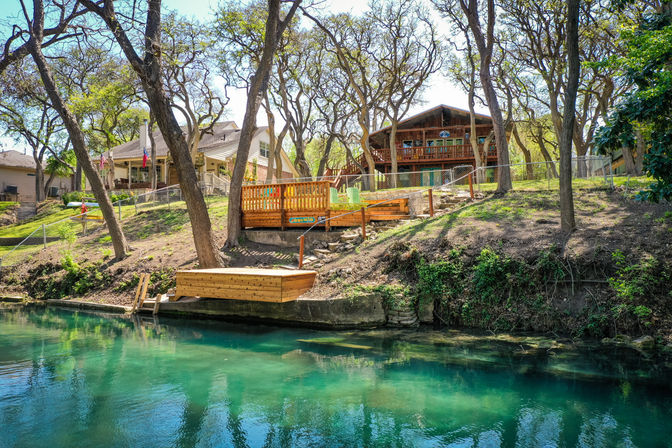 Sunny riverfront scene with clear turquoise water, wooden dock and raised deck with colorful Adirondack chairs, oak trees along the sloped shoreline and lakeside homes
