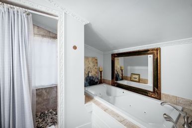 Attic-style bathroom with a white jetted soaking tub, marble surround, ornate gold-framed mirror, pebble-floor walk-in shower with white curtain, decorative crown molding and candle decor.