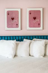Cozy modern bedroom scene with teal velvet headboard, four white pillows on a neatly made bed and two framed pink playing-card style 'Lucky' heart artworks on a pastel pink wall.