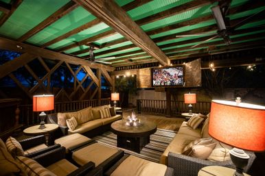 Cozy covered backyard patio with wicker sofas and ottomans, central fire pit, wall-mounted outdoor TV showing a game, warm table lamps and exposed wooden beams under a green ceiling — ideal outdoor living and entertaining space at dusk.