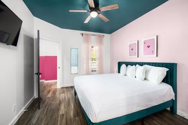 Stylish modern bedroom interior with teal ceiling and upholstered teal bed, pale pink accent wall, white bedding and pillows, wood-look flooring, ceiling fan, sheer window curtains, wall-mounted TV and open door revealing a pink hallway.