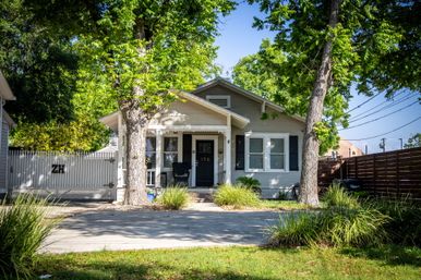 Sunlit gray bungalow with covered front porch and black door, flanked by two large shade trees, white picket fence, driveway and green lawn