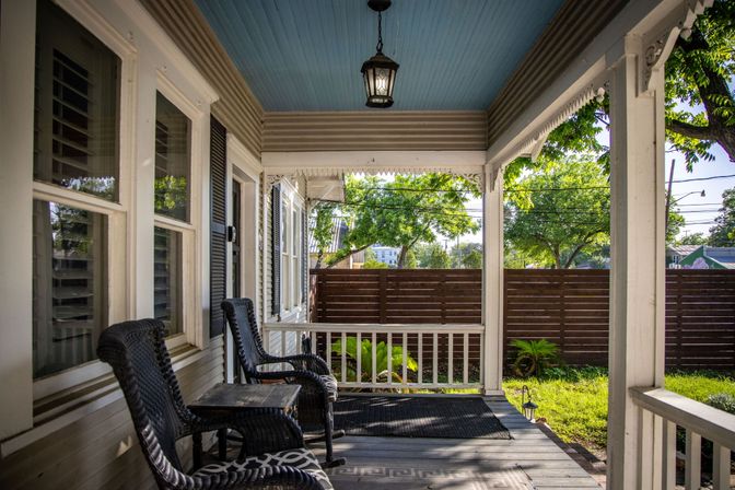 Cozy sunny front porch with blue beadboard ceiling, hanging lantern, two black wicker rocking chairs and small table overlooking a wooden privacy fence and leafy yard