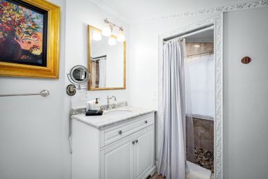 Bright white bathroom with marble countertop vanity, gold-framed mirror and floral painting, wall-mounted magnifying mirror, and a shower with light-gray curtain and pebble-tile floor.