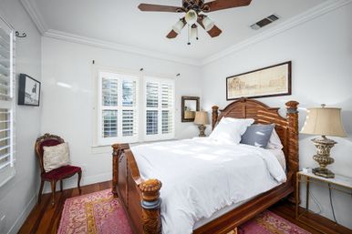 Sunlit bedroom with ornate carved wooden bed, crisp white bedding, plantation shutters, ceiling fan, vintage bedside lamps, red velvet chair and pink patterned rug.