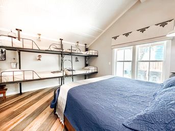 Bright cozy cottage bedroom with navy quilted queen bed in foreground, stacked black metal bunk beds along left wall, large double windows with roman shade, vaulted white beadboard ceiling and wide-plank wood floors.