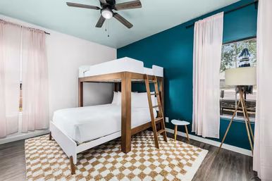 Sunlit modern bedroom with wooden bunk bed featuring lower bed and upper bunk, teal accent wall, pale pink curtains, patterned area rug, ceiling fan and tripod floor lamp