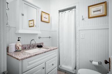 Bright white beadboard bathroom with speckled granite vanity, chrome faucet, mirrored medicine cabinet, gold-framed artwork and a white shower curtain — cozy small-bathroom design