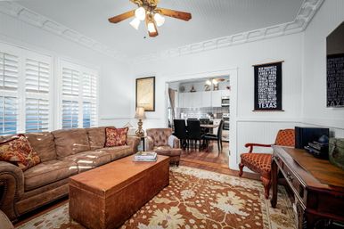 Sunny cozy living room with white plantation shutters, tufted brown sofa, leather trunk coffee table, patterned area rug, decorative crown molding and ceiling fan, opening to a dining area and kitchen.