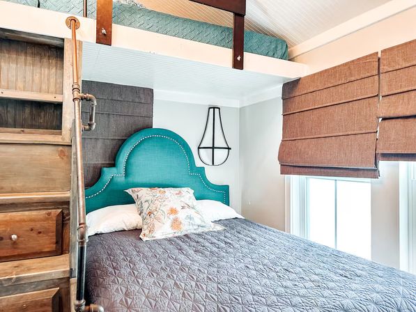 Cozy sunlit loft bedroom with a turquoise upholstered headboard, quilted gray bedding and floral pillow, wooden ladder with copper-pipe railing to an upper bunk, and brown Roman shades over windows.