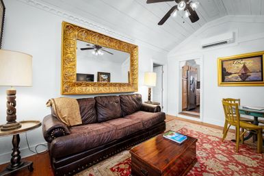 Cozy bright living room with a dark brown leather sofa, oversized gold ornate mirror reflecting a ceiling fan, wooden trunk coffee table on a red patterned rug, sailboat painting, small dining set, white shiplap ceiling and doorway leading to a kitchen.