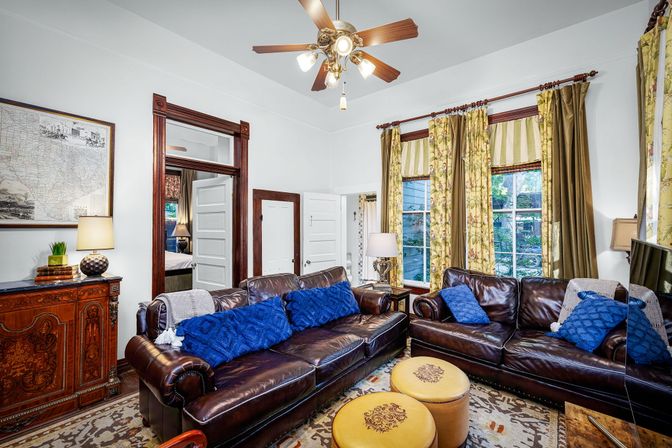 Cozy Victorian-style living room with dark leather sofas, bright blue throw pillows, yellow floral curtains over tall windows, round mustard ottomans, ornate wooden cabinet and brass ceiling fan