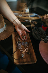 Hand wearing delicate bracelets reaches for boho necklaces and sparkly starburst earrings on a wooden tray next to a straw hat — cozy tabletop jewelry display.