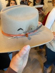 Hand-held tan felt fedora with an orange leather band, speckled feather and small mountain embroidery, photographed indoors with shoppers blurred in the background.
