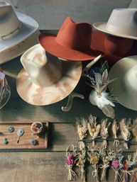 Top-down view of western-inspired wide-brimmed felt hats in cream, tan and rust with feather accents, hat pins, and tied dried flower bouquets arranged on a rustic wooden table.
