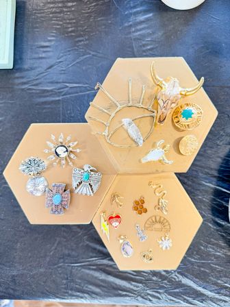 Boho southwestern-inspired jewelry display with gold and silver pins, turquoise stones, bull skull and sun eye charms arranged on honeycomb hexagon trays on a black table.