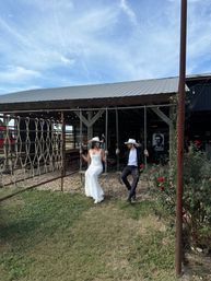 Rustic barn wedding scene: bride in a lace gown and groom in a suit wearing white cowboy hats sit on hanging swings beneath a wooden pavilion with string lights and red roses under a blue sky.