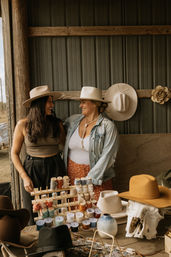 Two smiling women in wide-brimmed cowboy hats at a rustic barn market stall, surrounded by handmade hats, rows of colorful thread spools, beaded jewelry and a bleached animal skull on a wooden table.