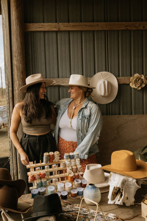 Two smiling women in wide-brimmed cowboy hats at a rustic barn market stall, surrounded by handmade hats, rows of colorful thread spools, beaded jewelry and a bleached animal skull on a wooden table.
