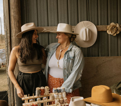 Two smiling women in wide-brimmed cowboy hats at a rustic barn market stall, surrounded by handmade hats, rows of colorful thread spools, beaded jewelry and a bleached animal skull on a wooden table.