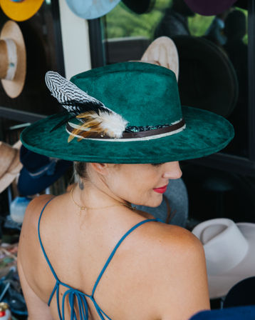 Woman in a blue strappy dress modeling a teal green wide-brim felt hat with feather accent in a hat shop/millinery display