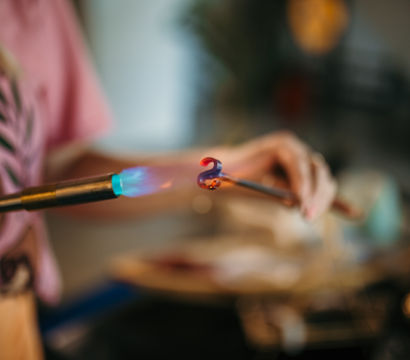 Close-up of blue torch flame heating a glass rod into a glowing red curled swirl in an artisan glassmaking studio workshop