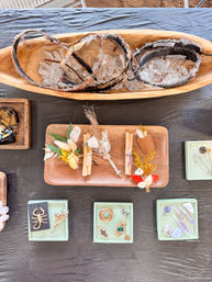 Overhead shot of a craft-fair table display: wooden bowls filled with quartz and feather headbands, wooden trays with dried flower boutonnieres, and small jewelry pieces on mint trays