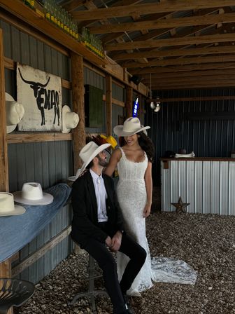Bride in lace gown and groom in black suit wearing white cowboy hats pose in a rustic Texas barn with wooden beams, hat display, corrugated metal walls and gravel floor.