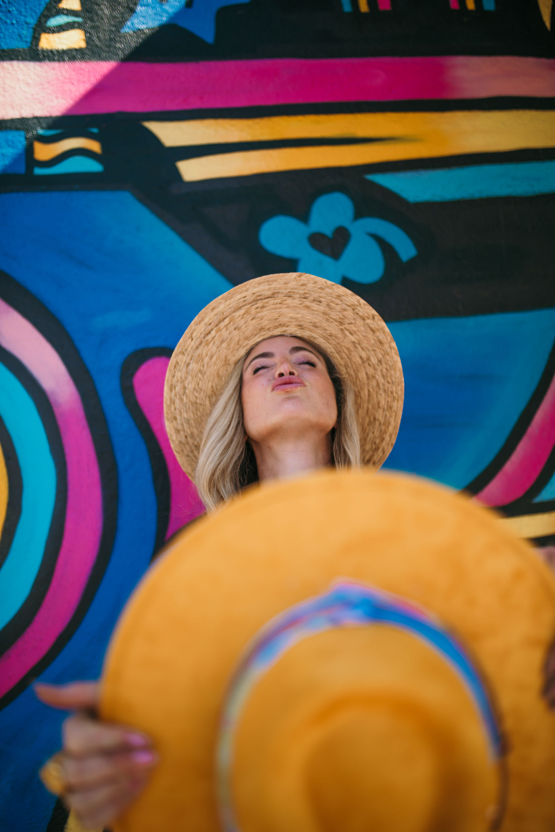 Person in a straw summer hat holding a bright yellow sunhat in front of a vibrant blue-and-pink street mural, playful puckered pose against colorful urban wall art