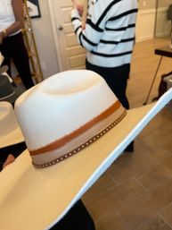 White felt wide-brim western cowboy hat with tan and rust ribbon and studded leather band, displayed indoors in a boutique with wooden floor and shoppers blurred in background