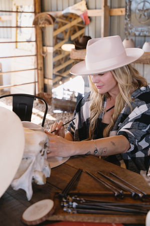 Woman artisan hand-shaping a pale pink wide-brim cowboy hat at a rustic western hat workshop, seated at a wooden table with hat blocks, shaping tools and a decorative skull