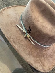 Top-down view of a tan suede cowboy hat with a thin studded leather band and small gold charm, resting on a rustic wooden table.