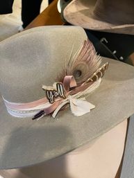 Tan wide-brim felt hat decorated with blush ribbons, peacock feather, studded trim and a small "y'all" pin, Southern-style accessory.