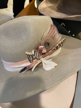 Tan wide-brim felt hat decorated with blush ribbons, peacock feather, studded trim and a small "y'all" pin, Southern-style accessory.