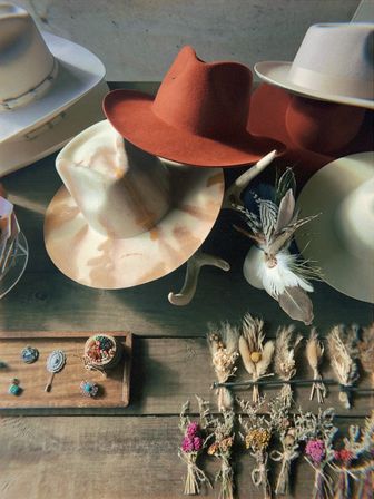 Rustic display of western felt hats (rust, cream, white) on an antler rack with feather accents, small jewelry and tied dried flower bouquets arranged on a wooden table