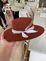 Stylish rust-colored felt wide-brim fedora with gray frayed ribbon, studded leather band, feather and crescent pin held over a marble kitchen island by rattan bistro chairs.