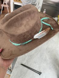 Brown suede cowboy-style hat with a turquoise ribbon and feather accent, held indoors over a light floor and wicker chair.