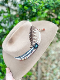 Tan felt cowboy hat with wide brim, patterned hatband, turquoise concho and striped feather, held against a blurred green garden background