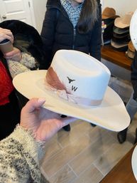 Hand holding a beige wide-brim felt western hat with a pink feather, satin ribbon trim and small longhorn emblem, displayed in a hat shop