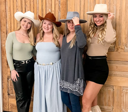 Four smiling women in cowgirl hats and western boots posing in ranch-inspired outfits in front of large rustic wooden paneled doors.