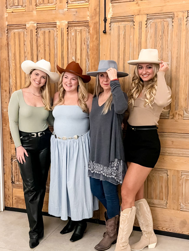 Four smiling women in cowgirl hats and western boots posing in ranch-inspired outfits in front of large rustic wooden paneled doors.