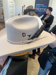 Beige felt western cowboy hat with suede band, silver buckle, black feather accent and small longhorn emblem with letter D, held indoors in a showroom setting.