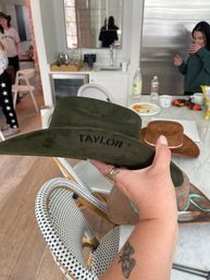 Green suede cowboy hat embroidered with "TAYLOR" held over a marble kitchen island, surrounded by other hats, drinks and wicker bistro chairs at a casual indoor gathering.