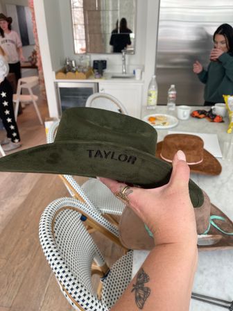 Green suede cowboy hat embroidered with "TAYLOR" held over a marble kitchen island, surrounded by other hats, drinks and wicker bistro chairs at a casual indoor gathering.