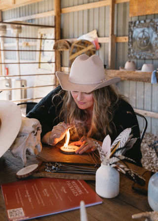 Person in a beige cowboy hat wood-burning a round slice in a rustic barn workshop, surrounded by a cow skull, feathers, tools and a wooden table.