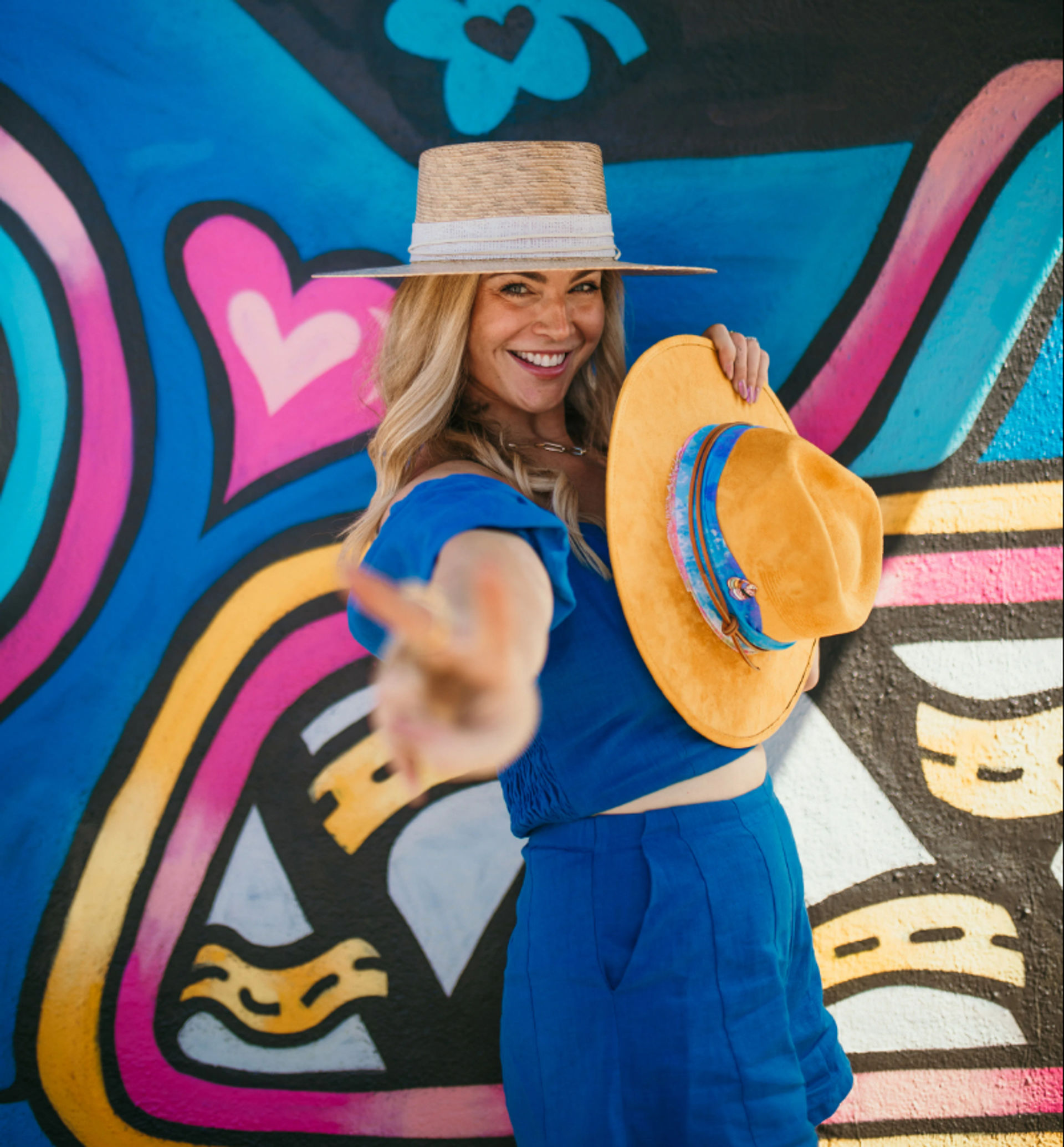Smiling woman in a blue outfit and straw hat holding a yellow hat, reaching toward the camera in front of a vibrant colorful street mural with hearts and abstract shapes