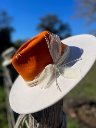 White wide-brimmed felt hat with burnt-orange crown and cream feather trim tied in a bow, perched on a weathered fence post against a bright blue sky in a sunny rural setting
