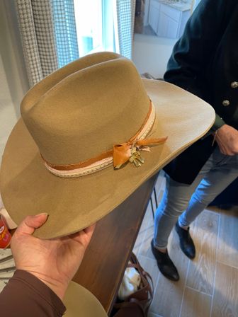 Hand holding a tan wide‑brim felt hat with a frayed leather ribbon and small charm, photographed indoors over a wooden table with someone in jeans and black ankle boots in the background.