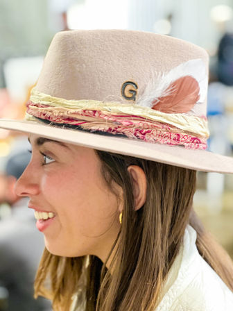 Side profile of a smiling person wearing a beige felt fedora with layered fabric bands, a white and peach feather, a stitched G initial, and a gold hoop earring against a blurred boutique interior.