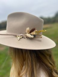 Boho-style beige wide-brim felt hat with dried floral accents and a small gold cow-skull charm on a ribbon hatband, worn over long blonde hair in a blurred grassy field